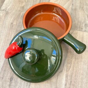 May include: A green ceramic casserole dish with a red chili pepper on the lid. The dish has a brown interior and a handle.