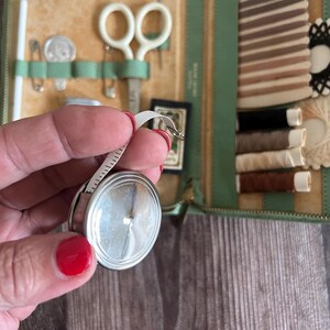 May include: A vintage sewing kit in a green leather case, open to reveal various sewing tools. Includes a silver tape measure, scissors, needles, thread, and a small card. The case is on a wooden surface.