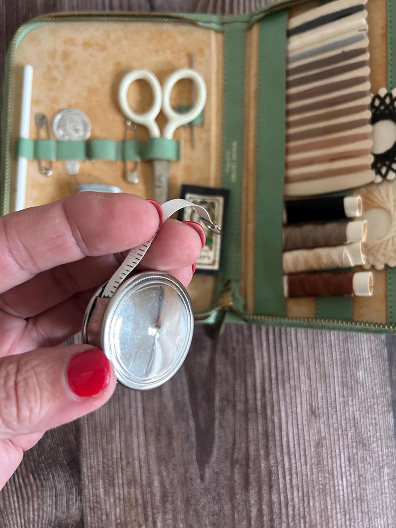 May include: A vintage sewing kit in a green leather case, open to reveal various sewing tools. Includes a silver tape measure, scissors, needles, thread, and a small card. The case is on a wooden surface.