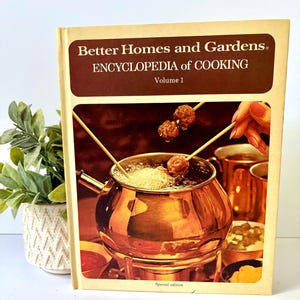 May include: A hardcover book titled Better Homes and Gardens Encyclopedia of Cooking Volume 1. The cover features a photograph of a copper fondue pot with skewers holding meatballs over bubbling oil, with the text Special edition at the bottom.