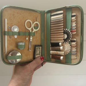 May include: Open green leatherette sewing kit with various sewing tools. The kit includes scissors, needles, spools of thread, and a small tape measure. The kit is open, revealing the contents, and is held by a person.