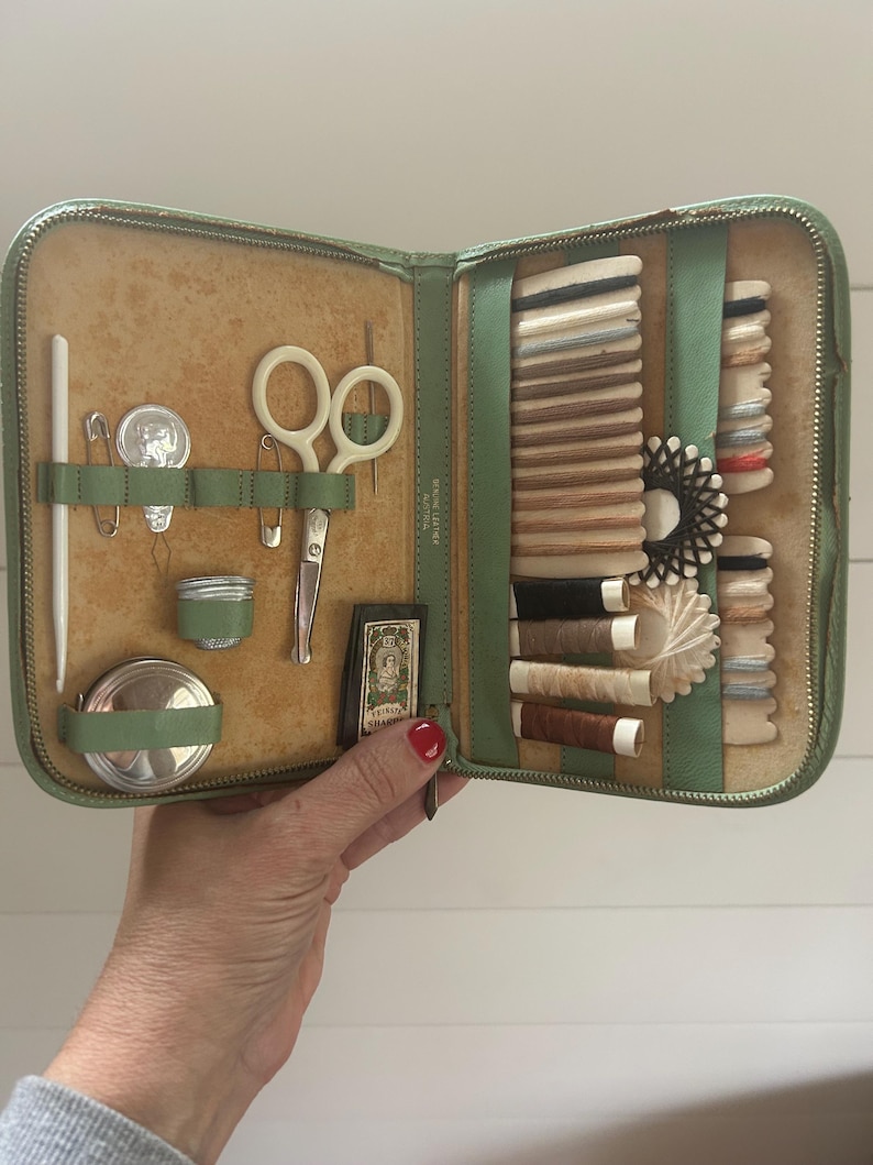 May include: Open green leatherette sewing kit with various sewing tools. The kit includes scissors, needles, spools of thread, and a small tape measure. The kit is open, revealing the contents, and is held by a person.