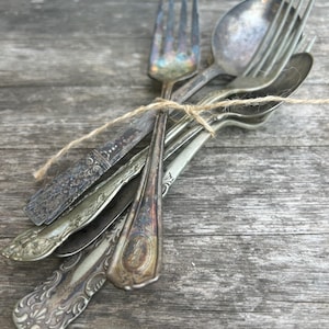 May include: A bundle of antique silverware, including forks, spoons, and knives, tied together with twine. The silverware has ornate, vintage designs and a tarnished, aged appearance, resting on a weathered wooden surface.