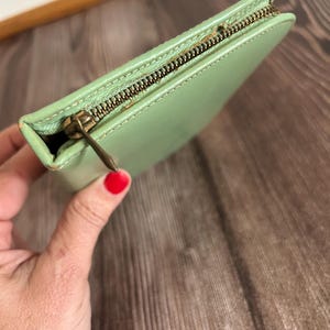 May include: A light green leather wallet with a brass zipper. The wallet is rectangular and appears to be closed. The zipper is a contrasting color to the wallet. The wallet is held in a hand.