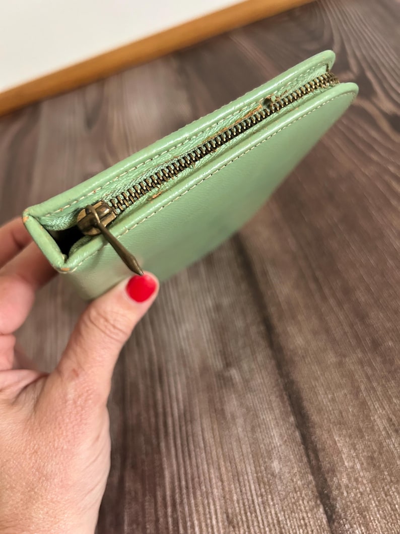May include: A light green leather wallet with a brass zipper. The wallet is rectangular and appears to be closed. The zipper is a contrasting color to the wallet. The wallet is held in a hand.