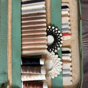 May include: Open vintage sewing kit with a light green leather exterior. The interior reveals spools of thread in various colors, including black, brown, and cream. Also visible are thread cards with multiple colors and a pair of scissors.