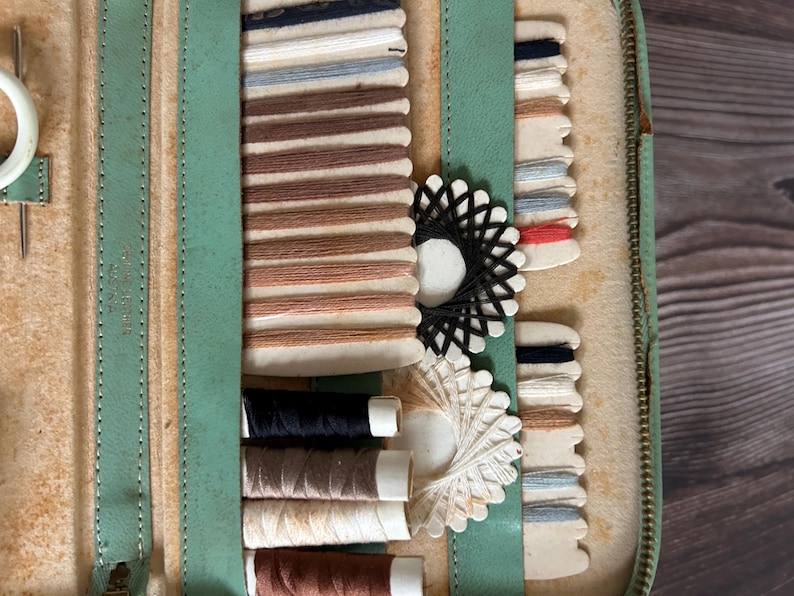 May include: Open vintage sewing kit with a light green leather exterior. The interior reveals spools of thread in various colors, including black, brown, and cream. Also visible are thread cards with multiple colors and a pair of scissors.