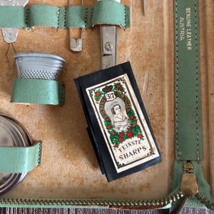 May include: An open vintage sewing kit with a variety of tools. The kit includes scissors, a thimble, a safety pin, and a packet of "Fineste Sharps" needles. The kit is made of tan leather with mint green accents and a zipper closure.
