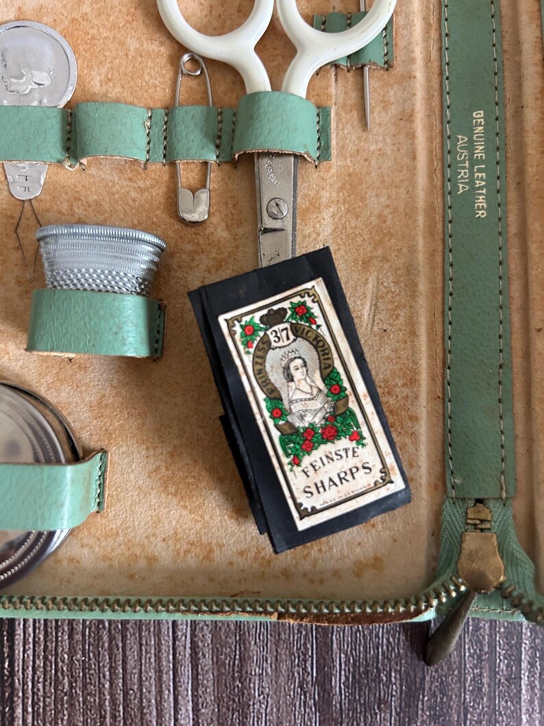 May include: An open vintage sewing kit with a variety of tools. The kit includes scissors, a thimble, a safety pin, and a packet of "Fineste Sharps" needles. The kit is made of tan leather with mint green accents and a zipper closure.