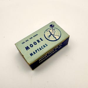 May include: A rectangular cardboard box for Moore Maptacks, featuring a light blue top with dark blue text reading 100-NO. 100 SERIES MOORE MAPTACKS and a globe graphic with a push pin. The side is dark blue with white text: MOORE PUSH PIN CO. PHILADELPHIA, PA.
