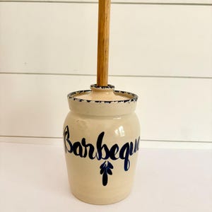 May include: A cream-colored ceramic crock with a lid and a wooden handle. The word "Barbeque" is written in dark blue cursive script on the front, accompanied by a small blue leaf icon. The rim of the lid features a dark blue speckled pattern.