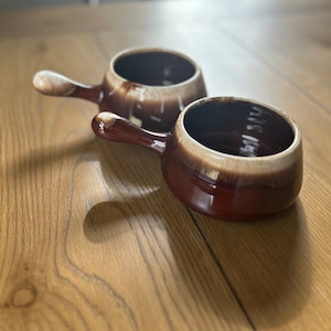 May include: Two brown and white ceramic soup bowls with handles. The bowls are on a wooden surface.