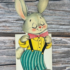 May include: Vintage Easter card featuring a cartoon rabbit with closed eyes and rosy cheeks. The rabbit wears a yellow shirt, green striped pants, and a red bow tie. Black suspenders and a fluffy tail complete the outfit. The card is set against a wooden background.