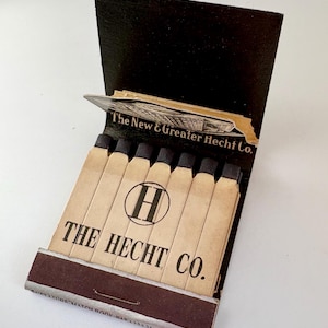 May include: A vintage cardboard matchbook with a black cover featuring the text The New & Greater Hecht Co. The interior contains six light tan matchsticks with black tips. The front displays a circular H logo and the text THE HECHT CO.