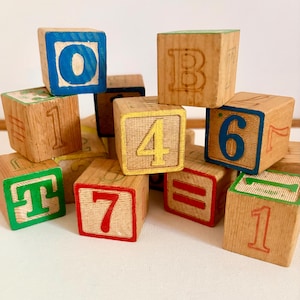 May include: A collection of wooden alphabet and number blocks stacked together. The blocks feature various colors including blue, green, red, and yellow, with printed characters such as the letters O, B, T and numbers 1, 4, 6, 7, and the equals sign.