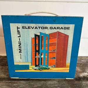 May include: A vintage blue metal toy carrying case with a white handle. The case features a colorful illustration of an elevator garage with the words "ELEVATOR GARAGE" and "MINI-LIFT". A green toy car is in front of the garage.