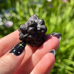 May include: A dark, textured, irregularly shaped stone, possibly a meteorite, held in a hand with a black manicure featuring a celestial design. The stone's surface appears rough and dark, contrasting with the skin tone and the green background.