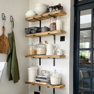 May include: A kitchen pantry with wooden shelves, black metal brackets, and various storage containers. The shelves hold bowls, jars, baskets, and canisters filled with dry goods. Two woven baskets sit on the floor, and a wooden cutting board and apron hang on the wall.