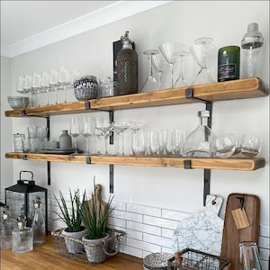 May include: A kitchen display with wooden shelves holding glassware, a cocktail shaker, and decorative items. Below, a countertop features plants in a wire basket, cutting boards, and bowls. The shelves are supported by black metal brackets.