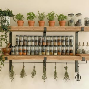 May include: A wooden spice rack with two shelves, holding glass jars of spices and herbs. Potted herbs sit on the top shelf. Below, a row of hanging dried herbs and a pair of scissors.