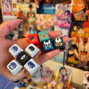 May include: A selection of square keycaps in various colours, each decorated with anime-style character designs. The keycaps are in red, blue, green, and white, with detailed illustrations. Some are attached to a keychain.
