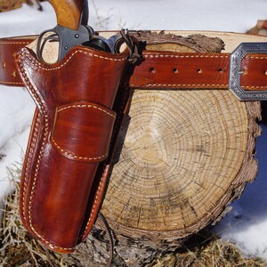 Könnte beinhalten: Ein braunes Lederholster mit einem Revolver, befestigt an einem passenden Gürtel mit einer silbernen Schnalle. Das Holster und der Gürtel sind mit weißem Faden genäht. Die Artikel liegen auf einem Baumstumpf.