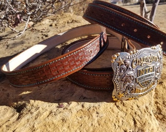 May include: A brown leather belt with a woven pattern and turquoise stitching. The belt has a silver and gold buckle with the text "Champion", "INTH", "NATIONAL TEAM ROPING", and a star.