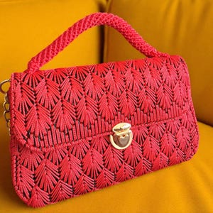 Op de afbeelding: Een rode, handgemaakte handtas met een geweven patroon en een goudkleurige sluiting. De tas heeft een gevlochten handvat en een kettingriem. De tas ligt op een gele ondergrond.