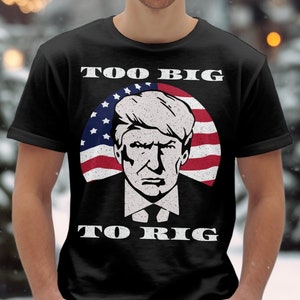 Puede incluir: Camiseta negra con un diseño de bandera estadounidense desgastado y un retrato de Donald Trump. El texto "Too Big To Rig" está impreso en la camiseta.