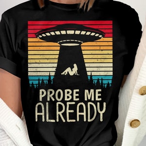 May include: Black t-shirt featuring a retro-style graphic of a UFO with a woman lying on the ground below it. The text "PROBE ME ALREADY" is printed below the graphic.