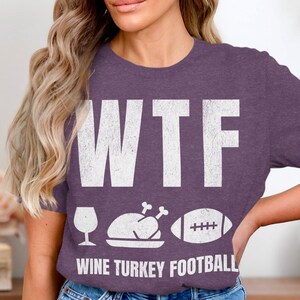 Può includere: Maglietta viola con testo bianco che recita "WTF WINE TURKEY FOOTBALL" con un bicchiere di vino, un tacchino e un pallone da calcio.
