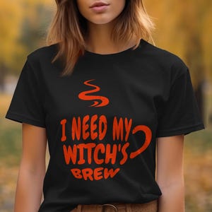 Op de afbeelding: Zwart T-shirt met een rode afbeelding van een koffiekopje met stoom en de tekst "Ik heb mijn heksendrank nodig".