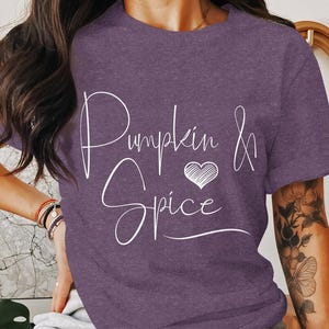 以下が含まれることがあります： 白い文字で「Pumpkin & Spice」と書いてある紫色のTシャツ。ハートのデザインが施されています。