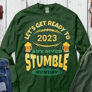 May include: Green long-sleeve t-shirt with a football graphic and the text "Let's Get Ready To 2023 Any Given Stumble Sunday".