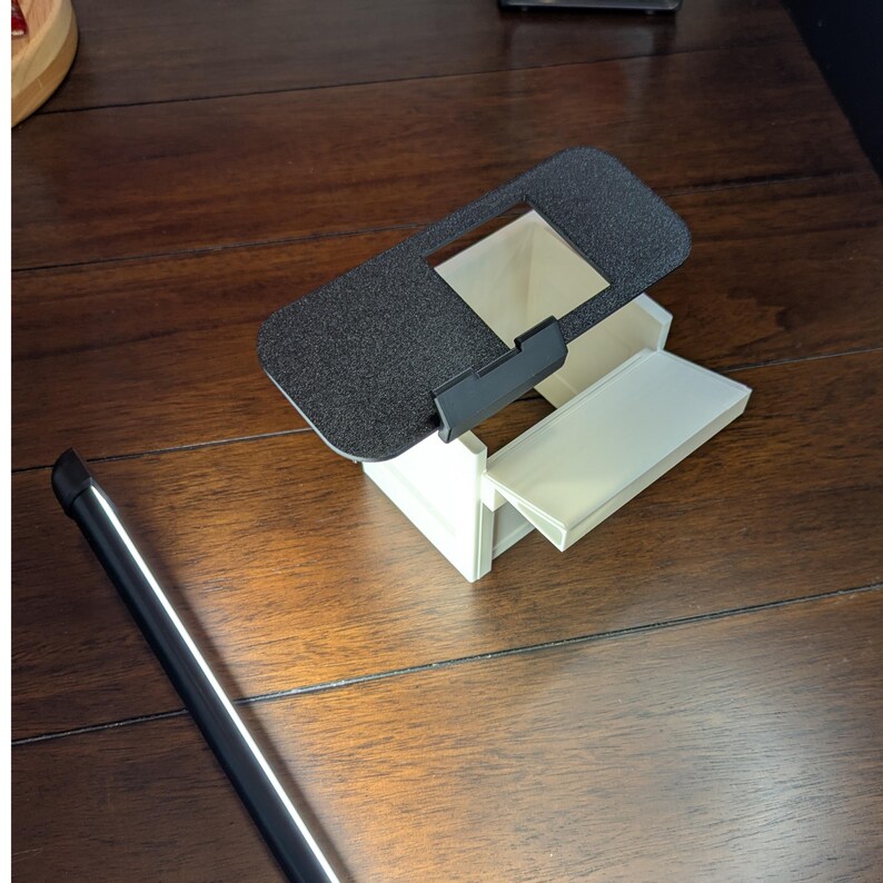 K&ouml;nnte beinhalten: Ein wei&szlig;es 3D-gedrucktes Modell eines kleinen Hauses mit einem schwarzen Deckel, der sich &ouml;ffnen l&auml;sst. Der Deckel hat eine strukturierte Oberfl&auml;che und eine rechteckige &Ouml;ffnung.