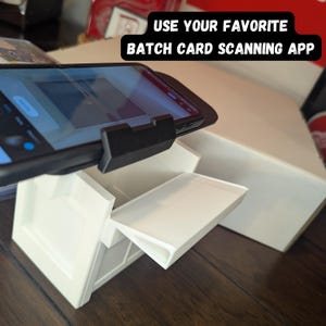 K&ouml;nnte beinhalten: Wei&szlig;e Kartenscan-Station mit einem Smartphone in einem schwarzen Etui, das oben befestigt ist. Das Telefon zeigt eine Kartenscan-App an. Der Text auf der wei&szlig;en Box lautet "USE YOUR FAVORITE BATCH CARD SCANNING APP".