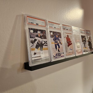 May include: A black shelf displays several hockey trading cards in protective cases. The cards feature players in action, with team logos and player names visible. The shelf is mounted on a light-colored wall.