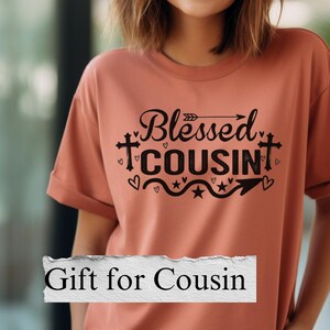 May include: A coral-colored t-shirt with the words "Blessed Cousin" in black script and a decorative design. The shirt also has the text "Gift for Cousin" in a white rectangle.
