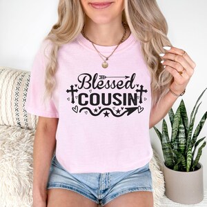 May include: A light pink t-shirt with the words "Blessed Cousin" in black script and a decorative arrow, hearts, and stars. The shirt is a crew neck and has short sleeves.