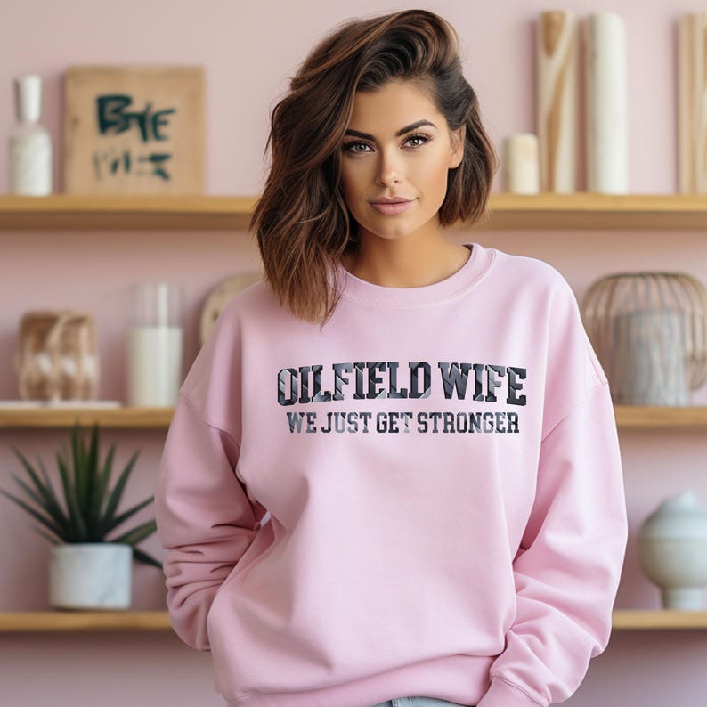 May include: A light pink sweatshirt with the text "OILFIELD WIFE WE JUST GET STRONGER" in a black and gray design. The sweatshirt is a crew neck style.