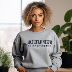 May include: A light gray sweatshirt with the text "OILFIELD WIFE" in a camouflage-style font. Below the text is the phrase "WE JUST GET STRONGER" in a simple font. The sweatshirt has long sleeves and a crew neck.