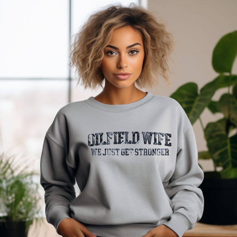 May include: A light gray sweatshirt with the text "OILFIELD WIFE" in a camouflage-style font. Below the text is the phrase "WE JUST GET STRONGER" in a simple font. The sweatshirt has long sleeves and a crew neck.