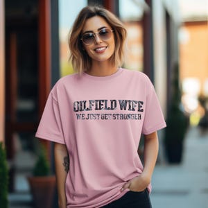 May include: A light pink t-shirt with the text "OILFIELD WIFE" in a patterned font above the text "WE JUST GET STRONGER" in a black font. The shirt has short sleeves.