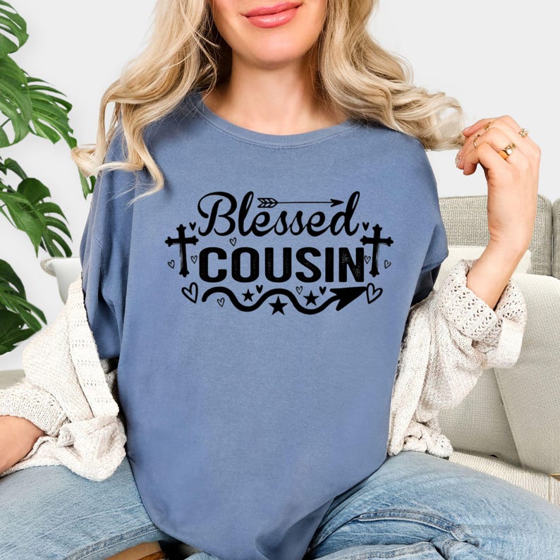 May include: A blue t-shirt with the words "Blessed Cousin" in black script and decorative elements. The design includes arrows, hearts, stars, and crosses. The shirt is a solid color and has a crew neck.