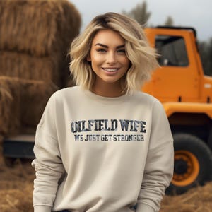 May include: A beige sweatshirt with the text "OILFIELD WIFE WE JUST GET STRONGER" in a dark blue and gray design. The image shows a woman smiling in front of a hay bale and an orange tractor.