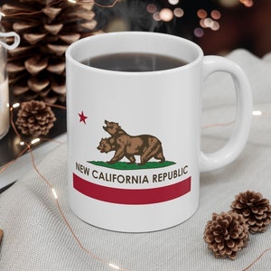 May include: White ceramic mug featuring the New California Republic flag design. The design includes a brown bear, a red star, and the text "NEW CALIFORNIA REPUBLIC" above a red stripe. The mug is filled with a dark liquid.