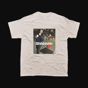 Op de afbeelding: Een lichtbeige t-shirt met een meme-afbeelding van een persoon die een beker vasthoudt, met het woord "Shhhhhh" eronder gedrukt. De afbeelding is vierkant, tegen een donkere achtergrond.