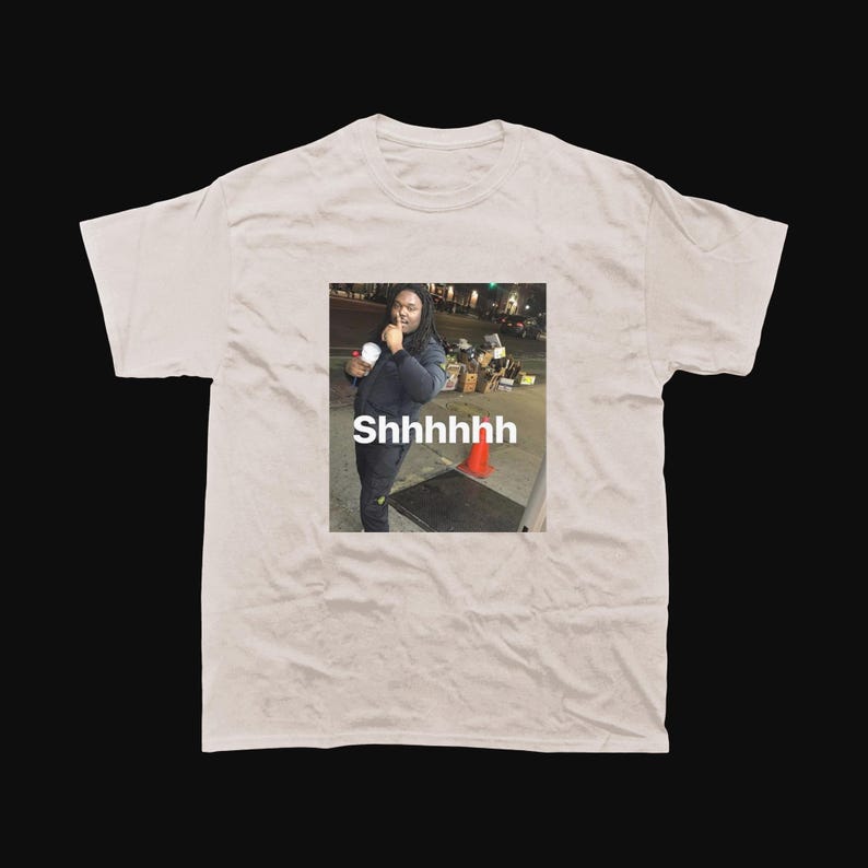Op de afbeelding: Een lichtbeige t-shirt met een meme-afbeelding van een persoon die een beker vasthoudt, met het woord "Shhhhhh" eronder gedrukt. De afbeelding is vierkant, tegen een donkere achtergrond.