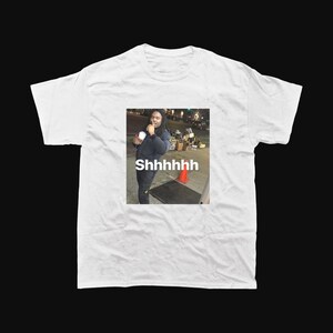 Op de afbeelding: Wit T-shirt met een meme van een persoon die een beker vasthoudt en met zijn vinger naar zijn lippen gebaart. De afbeelding op het shirt heeft het woord "Shhhhhhh" eronder gedrukt.