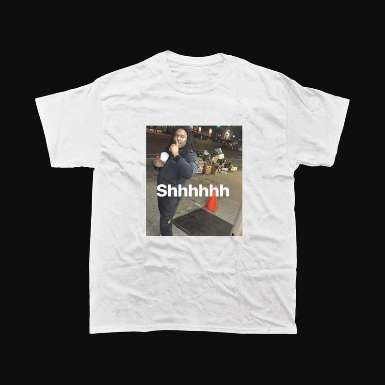 Op de afbeelding: Wit T-shirt met een meme van een persoon die een beker vasthoudt en met zijn vinger naar zijn lippen gebaart. De afbeelding op het shirt heeft het woord "Shhhhhhh" eronder gedrukt.